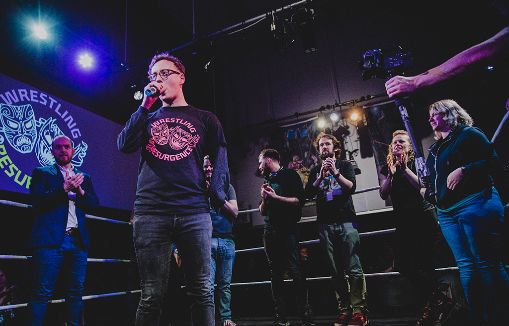 Sam West speaks to a Wrestling Resurgence audience from inside the wrestling ring as performers and collaborators applaud.