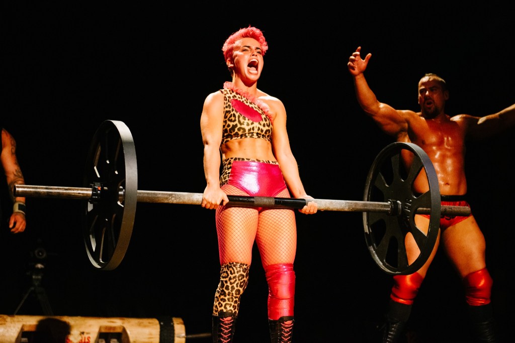 Clementine lifts a barbell onstage while Charles Crowley cheers behind her during Unruly Music Hall Sport.